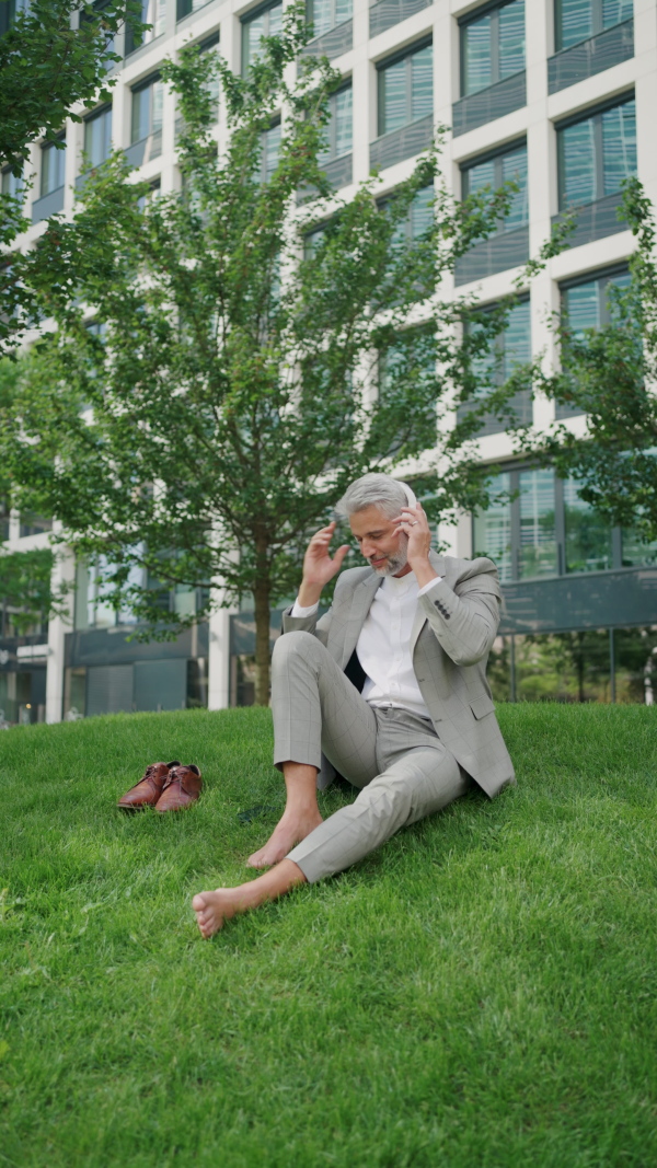 Mature businessman sitting barefoot on grass outside office building, using smart phone, listening to music and relaxing.