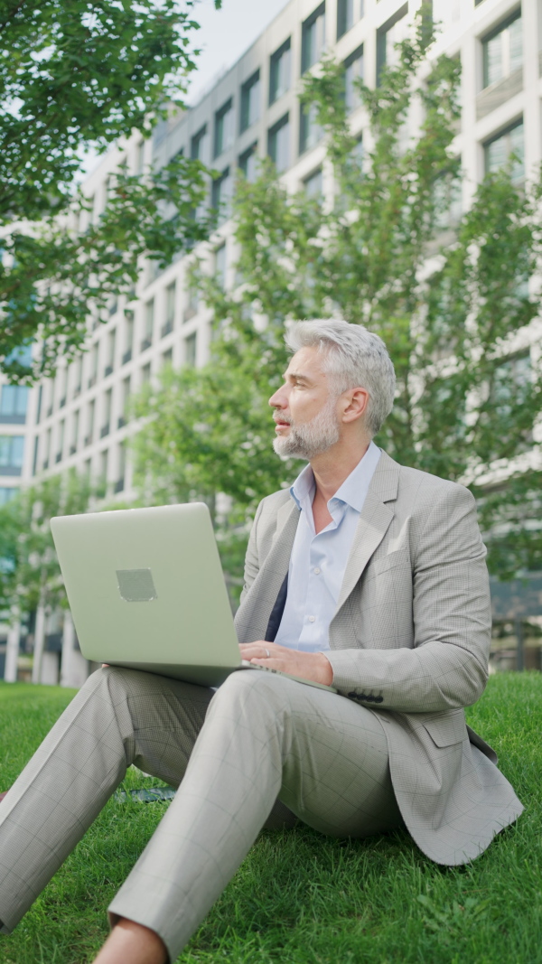 Mature businessman working on laptop in front of office building, vertical footage.