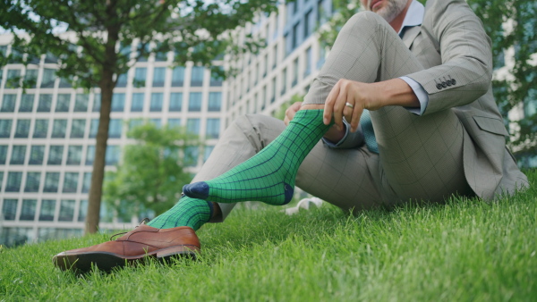 Mature businessman putting on shoes and socks on grass in front of office building.