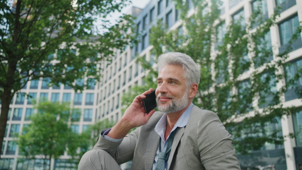 Mature businessman sitting outside office building after work, ,talking on phone with loosed tie. Green environment. Close up.