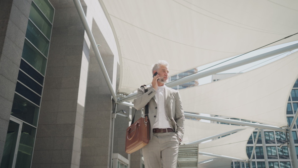 Mature businessman walking outside office building using smart phone in city, talking and smiling. Low angle view.