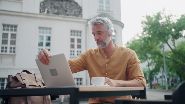 Man working on laptop in outside coffee shop, wearing headphones.