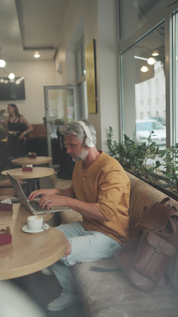 Mature male commuter sitting in cafeteria, working on laptop, using phone and wearing headphones.