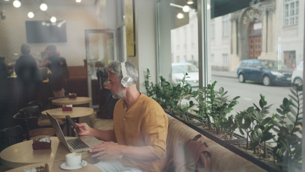 Man working on laptop inside coffeeshop, wearing headphones. Window shot.