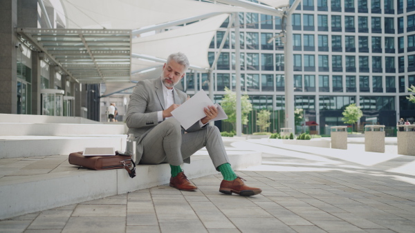 Mature businessman sitting outside office building using smart phone , thinking.