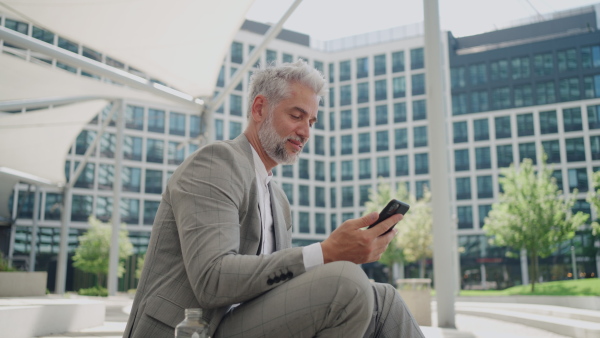 Mature businessman sitting outside office building using smart phone , thinking.