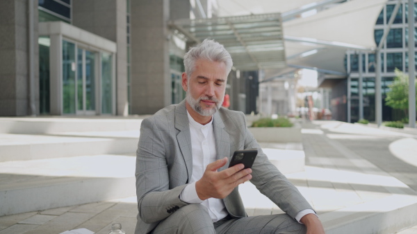 Mature businessman sitting outside office building using smart phone ,looking at camera.