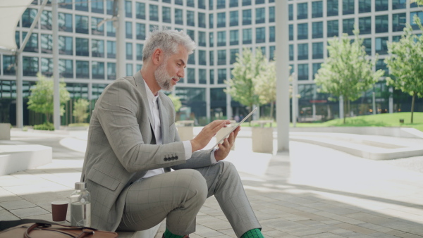 Mature businessman sitting outside office building using tablet in city during break. Medium shot.