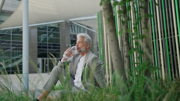 Mature businessman sitting outside office building during lunch break, drinking water, being content. Medium full shot.