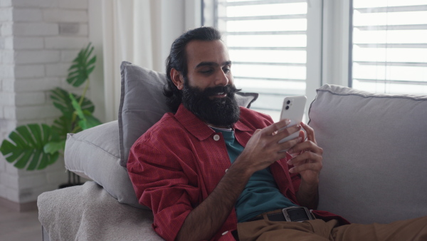 A man sitting on sofa, scrolling on smartphone in living room. Video of handsome indian man, phone in hand, smiling.