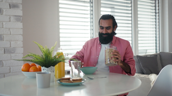 A man sitting in kitchen, preparing breakfast, smiling. Video of handsome indian man, having a bowl of cereals, smiling.