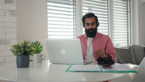 A man working from home office on laptop, making phone call with bluetooth headset. Handsome Indian man in coat, working remotely from apartment. Student at online class.