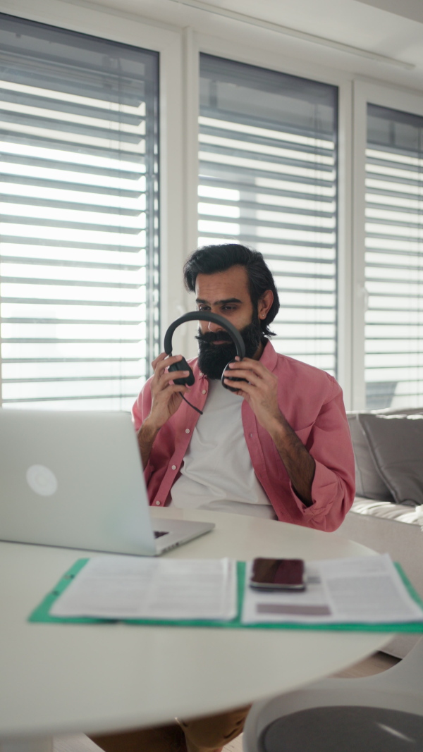 A man working from home office on laptop, making phone call with bluetooth headset. Handsome Indian man in coat, working remotely from apartment. Student at online class. Vertical view.