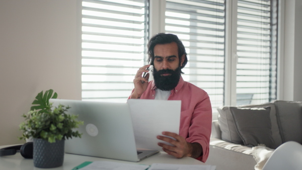 A man working from home office on laptop, making phone call. Handsome Indian man in coat, working remotely from apartment.