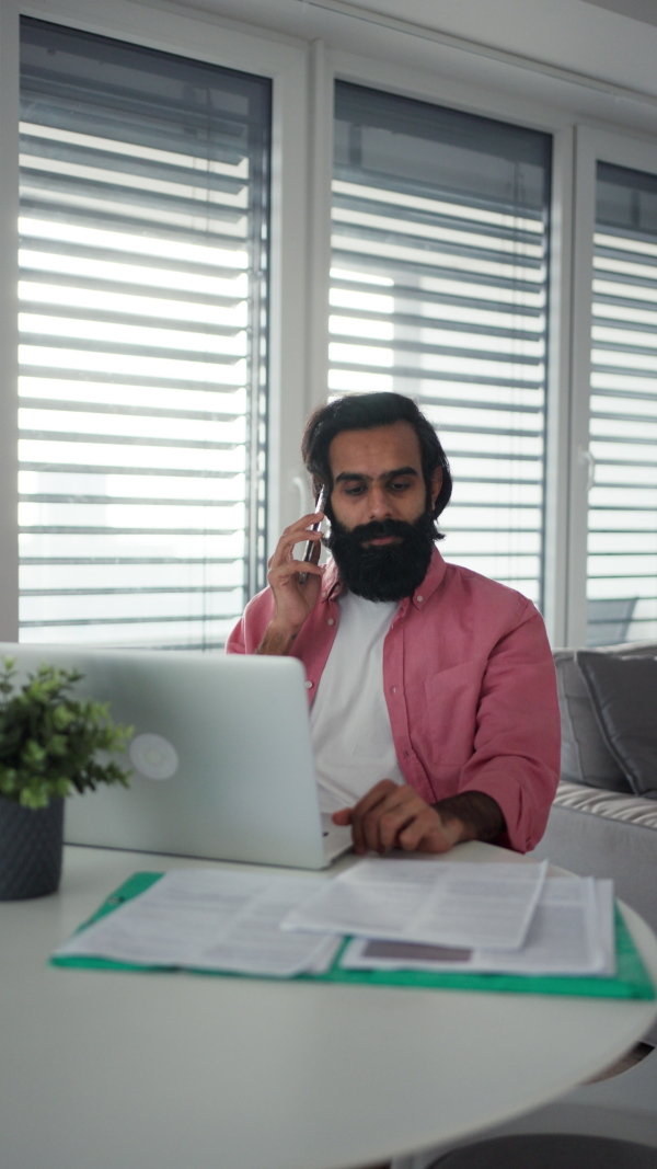 A man working from home office on laptop, making phone call. Handsome Indian man in coat, working remotely from apartment. Vertical view.