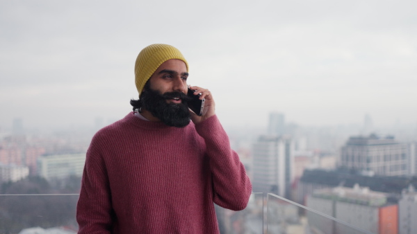 A man standing on terrace,having a call. Handsome indian man at home, in pink sweater and orange knit hat.