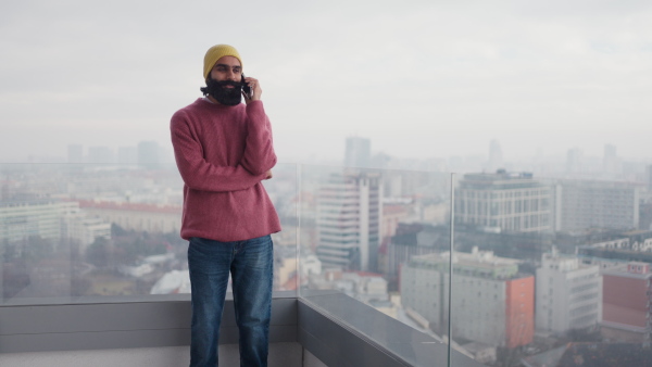 A man standing on terrace,having a call. Handsome indian man at home, in pink sweater and orange knit hat.