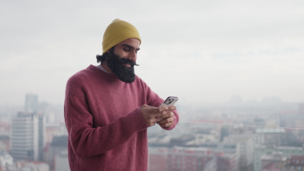 A man standing on terrace, scrolling on smartphone. Handsome indian man at home, in pink sweater and orange knit hat.