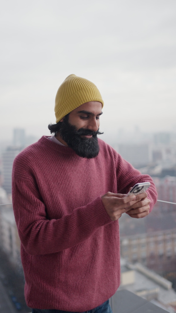 A man standing on terrace, scrolling on smartphone. Handsome indian man at home, in pink sweater and orange knit hat. Vertical view.