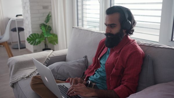 A man working from home, home office from kitchen, working on laptop, holding smartphone. Handsome Indian man in pink shirt, working remotely from apartment.