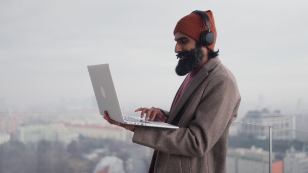 A man working from terrace, home office, working on laptop, making phone call with bluetooth headset. Handsome Indian man in coat, working remotely from outdoor. Student at online class.