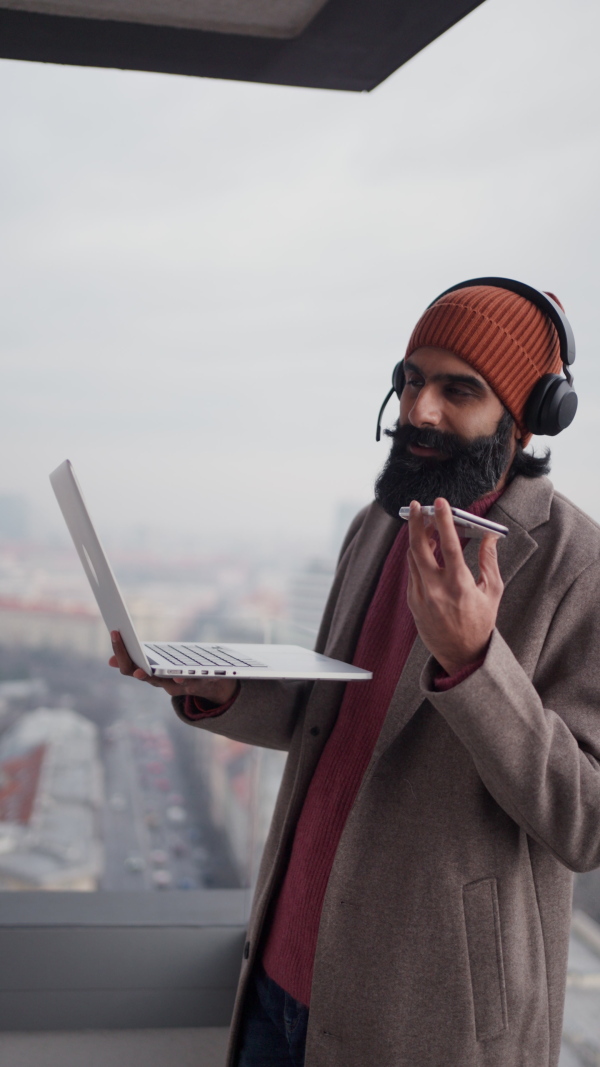 A man working from terrace, home office, working on laptop, making phone call with bluetooth headset. Handsome Indian man in coat, working remotely from outdoor. Student at online class. Vertical view.