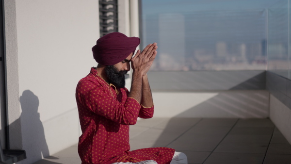 A man sitting on terrace, praying, meditating at home in the morning. Video of handsome Indian man with turban.