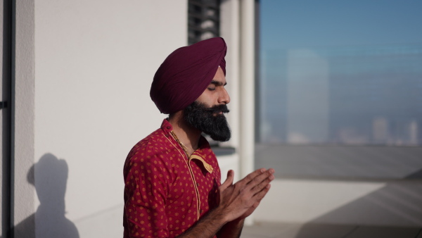 A man sitting on terrace, praying, meditating at home in the morning. Video of handsome Indian man with turban.