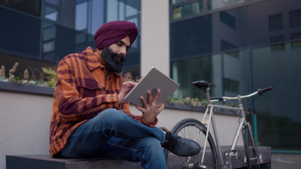 An young Indian man phone calling, sitting in front of office building, working on tablet. Video of handsome Indian man with turban, during cold spring day.