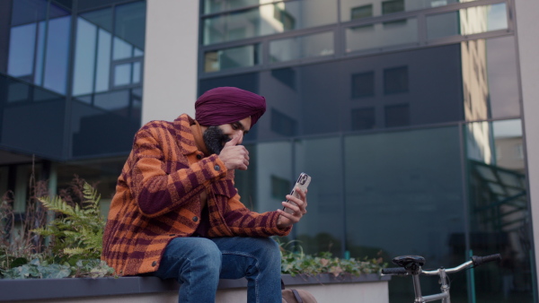 A video of Indian man phone calling, sitting in front of office building, smiling. Portrait of handsome Indian man with turban, during cold spring day.