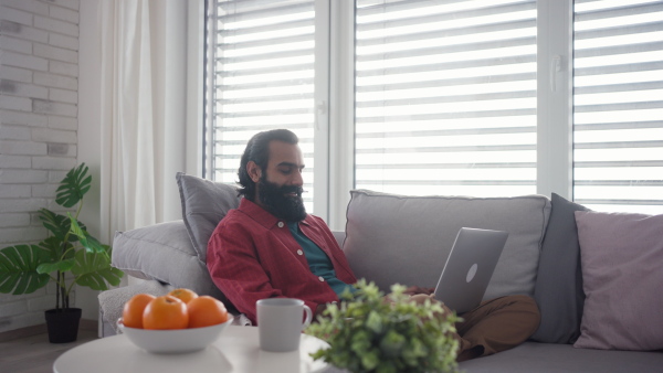 A man working from home, home office from kitchen, working on laptop, holding smartphone. Handsome Indian man in pink shirt, working remotely from apartment.