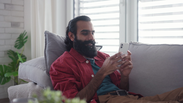 A man sitting on sofa, scrolling on smartphone in living room. Video of handsome indian man, phone in hand, smiling.