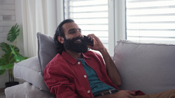 A man sitting on sofa,having a call in living room. Video of handsome indian man, phone in hand, smiling.