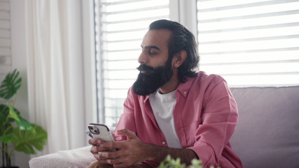 A man sitting on sofa, scrolling on smartphone in living room. Video of handsome indian man, phone in hand, smiling.