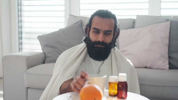 A man is sick, drinking hot herbal tea and taking medication, painkillers. Handsome indian man at home with cold.