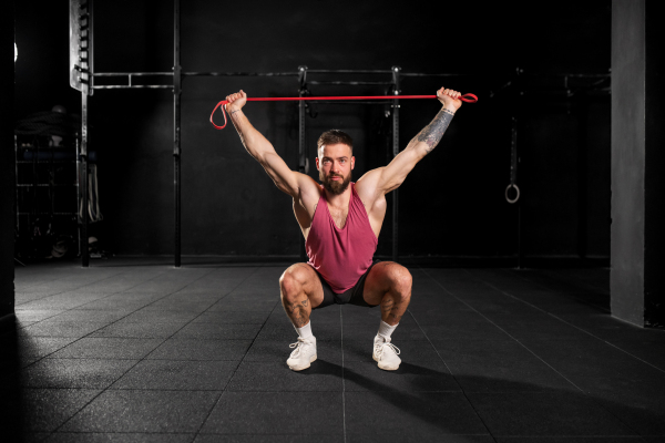 Portrait of handsome muscular man training with resistance band in gym, squatting. Routine workout for physical and mental health.