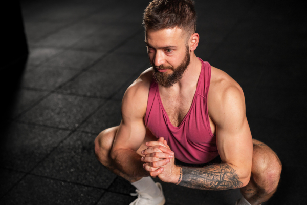 Portrait of handsome muscular man stretching his body, squatting, in gym. Routine workout for physical and mental health.