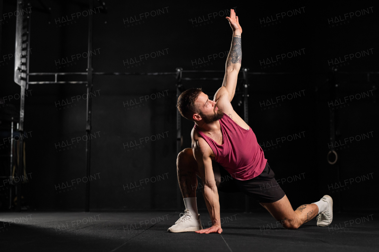 Portrait of handsome muscular man stretching his body, squatting, in gym. Routine workout for physical and mental health.