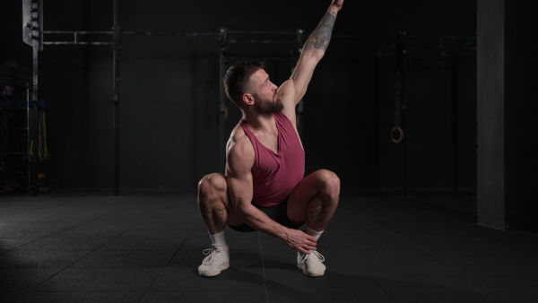 Video of handsome muscular man stretching his body, squatting, in gym. Routine workout for physical and mental health.