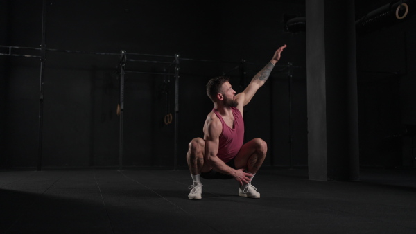 Video of handsome muscular man stretching his body, squatting, in gym. Routine workout for physical and mental health.