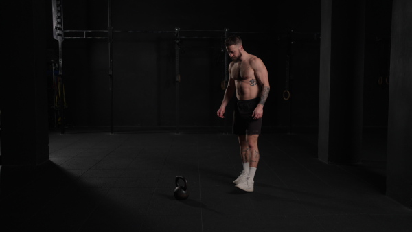 Man lifting dumbbell from gym floor, wearing only shorts, bare chest. Routine workout for physical and mental health.