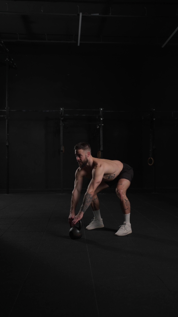 Man lifting dumbbell from gym floor, wearing only shorts, bare chest. Routine workout for physical and mental health.