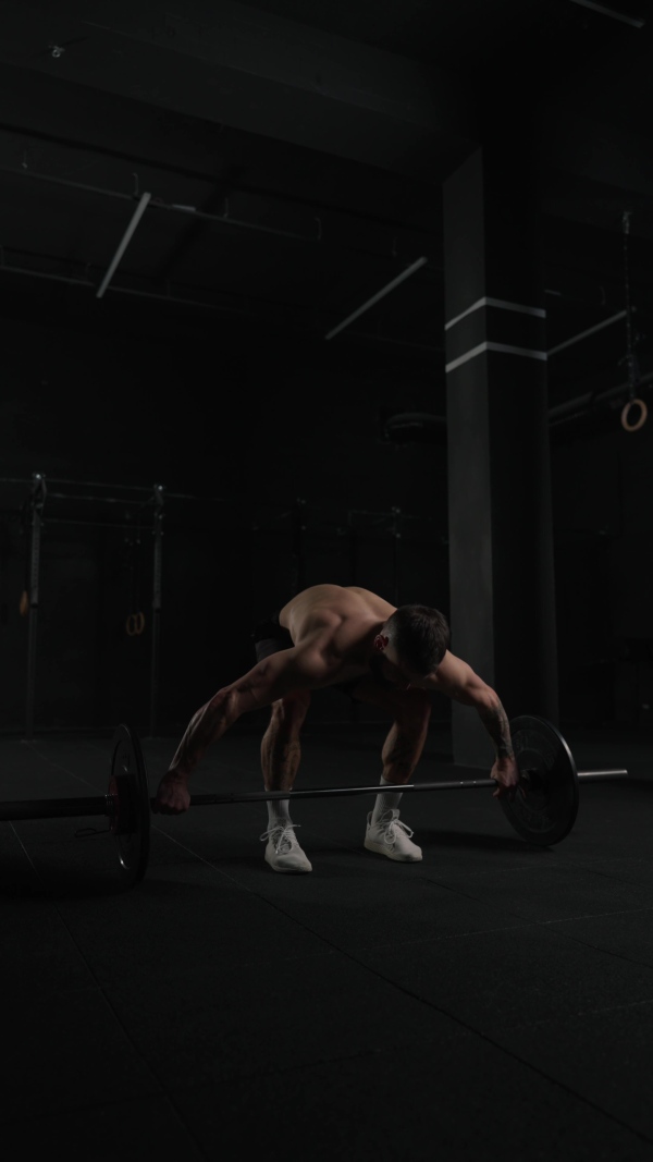 Man lifting dumbbell from gym floor, wearing only shorts, bare chest. Routine workout for physical and mental health.