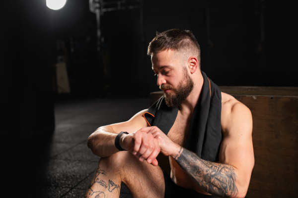 Handsome muscular man checking his performance on smartwatch, resting after a training in gym. Routine workout for physical and mental health.