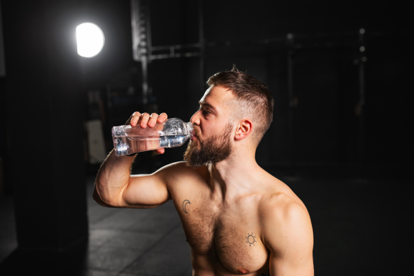 Muscular man resting after exercise, drinking water from bottle, sitting, wearing short with muscular bare chest. Routine workout for physical and mental health.