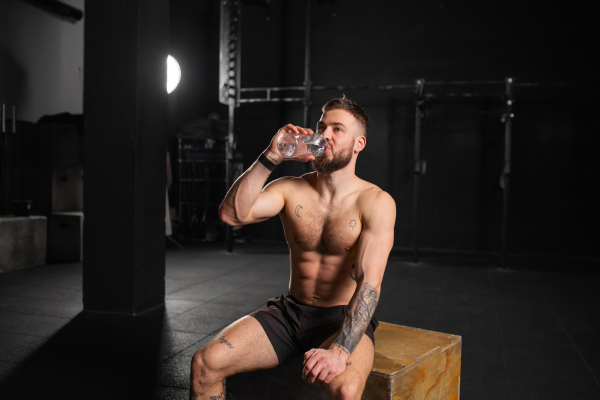 Muscular man resting after exercise, drinking water from bottle, sitting, wearing short with muscular bare chest. Routine workout for physical and mental health.