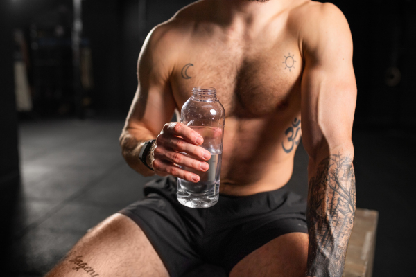 Muscular man resting after exercise, drinking water from bottle, sitting, wearing short with muscular bare chest. Routine workout for physical and mental health.