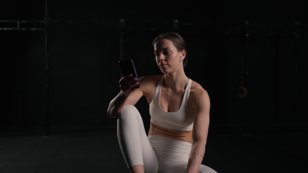 Fit young woman resting after training in gym, scrolling on smartphone, checking workout app. Routine workout for physical and mental health.