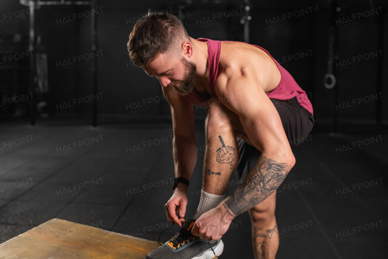 Handsome muscular man tying shoelaces on his sneakers in gym. Routine workout for physical and mental health.