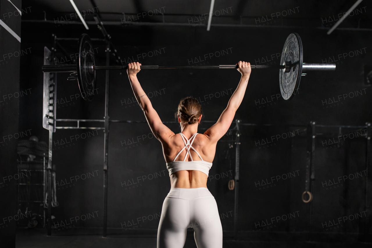 Rear view of strong woman performing overhead squat, pressing barbell up, standing up from squating. Routine workout for physical and mental health.